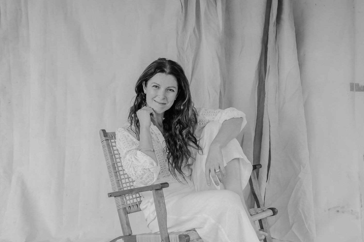 Black and white portrait of a smiling brunette woman in a white dress sitting in a wooden chair with draped fabric backdrop.