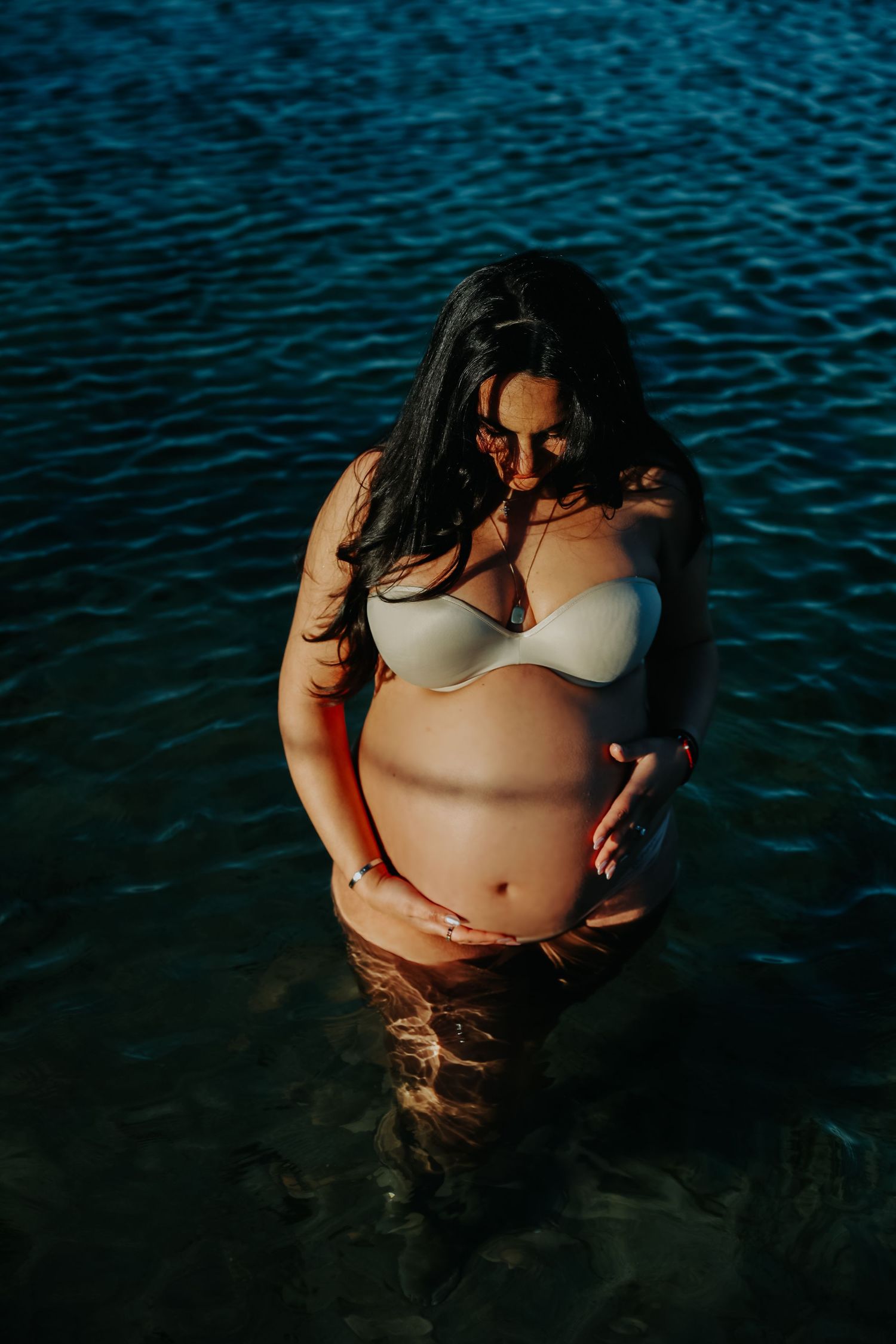 Pregnant woman in white bikini cradling baby bump while standing in dark blue water.