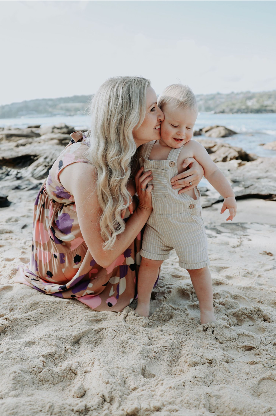 Blonde mother kneeling on sandy beach kissing baby boy in linen overalls near rocky shoreline.