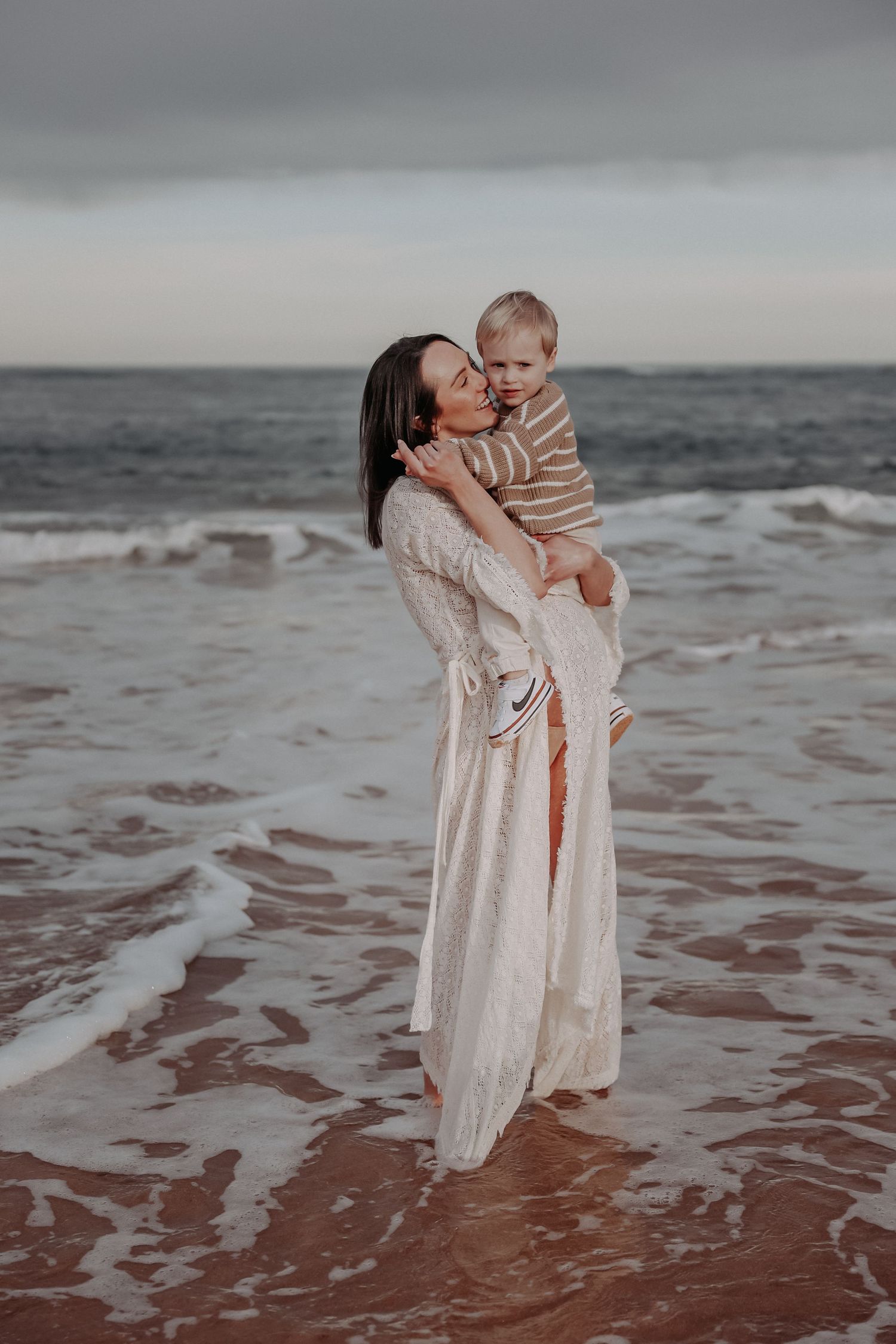 A mother in a white lace dress holds her toddler on a beach with ocean waves at dusk.