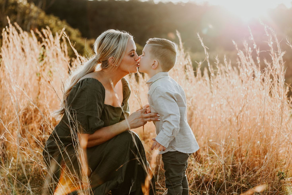 A pregnant mother shares a tender kiss with her young son in a sunlit golden grass field.