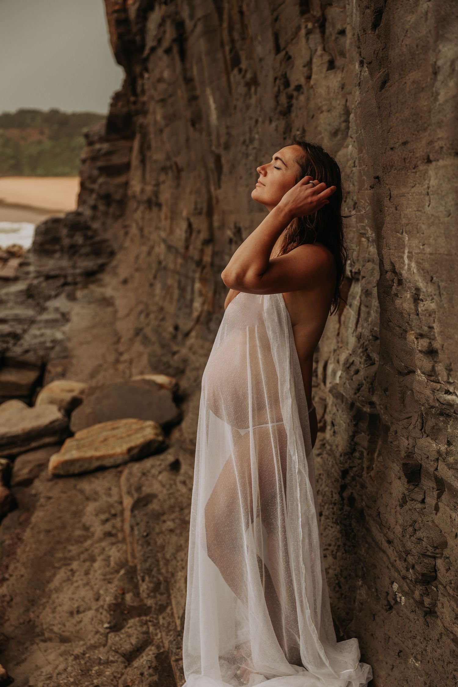Woman in sheer white dress leaning against rocky cliff wall at beach, hands raised to hair.