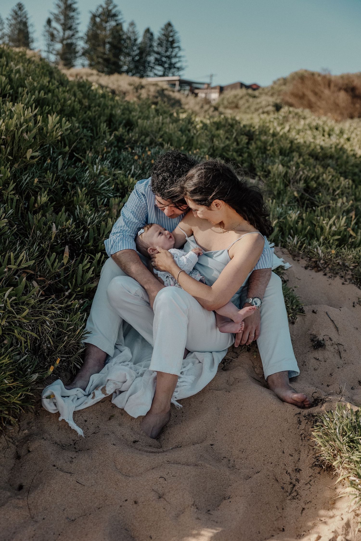 A couple sits barefoot on sandy beach dunes cradling their newborn baby, surrounded by lush green coastal vegetation.