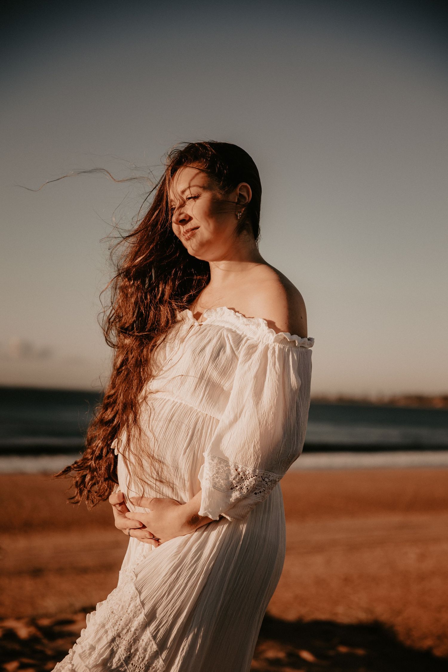 Pregnant woman in white off-shoulder dress on beach, hair blowing in wind at golden hour.