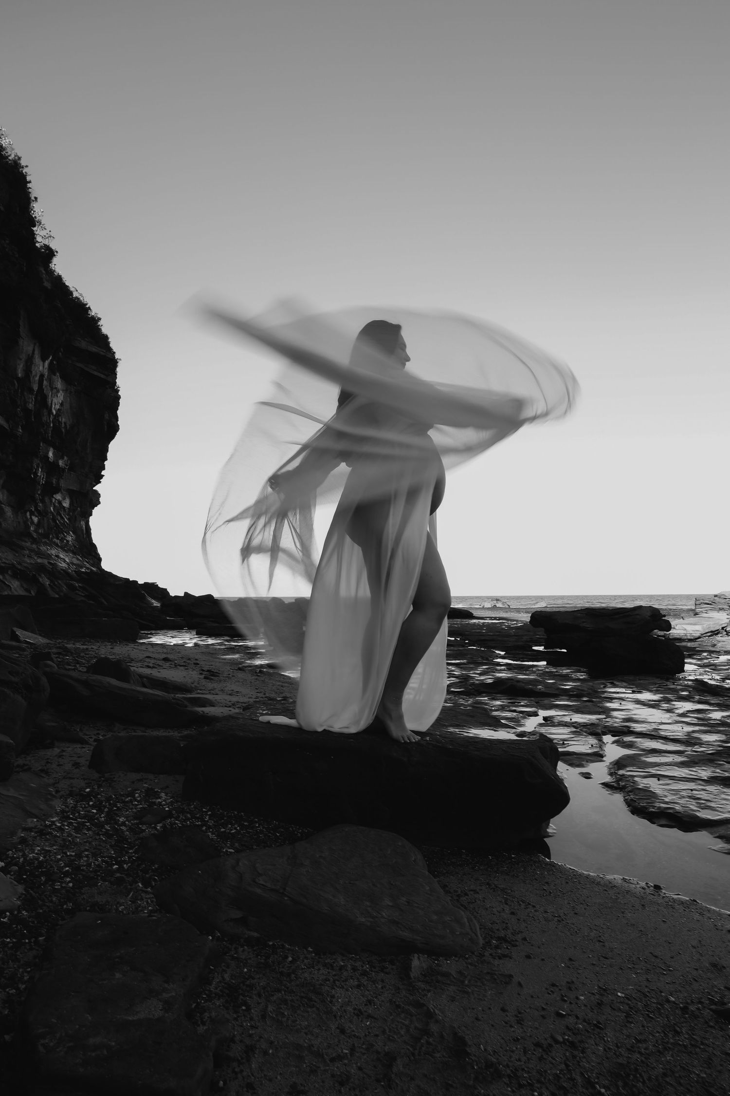 Black and white photo of a woman in flowing white dress dancing on rocky beach with motion blur effect.
