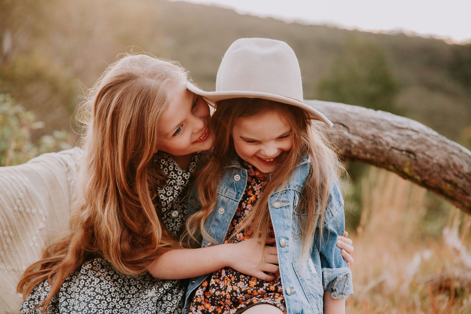 Two young girls with long hair laughing and hugging in a sunlit meadow.