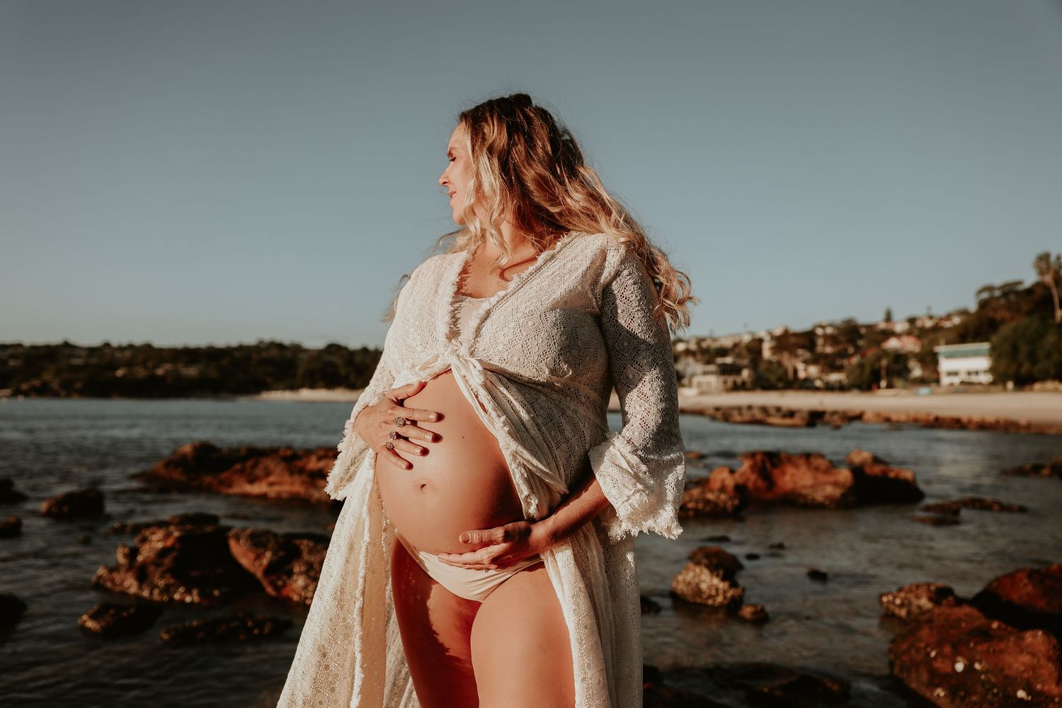 Pregnant woman in white lace robe cradling baby bump at rocky beach during golden hour sunset.