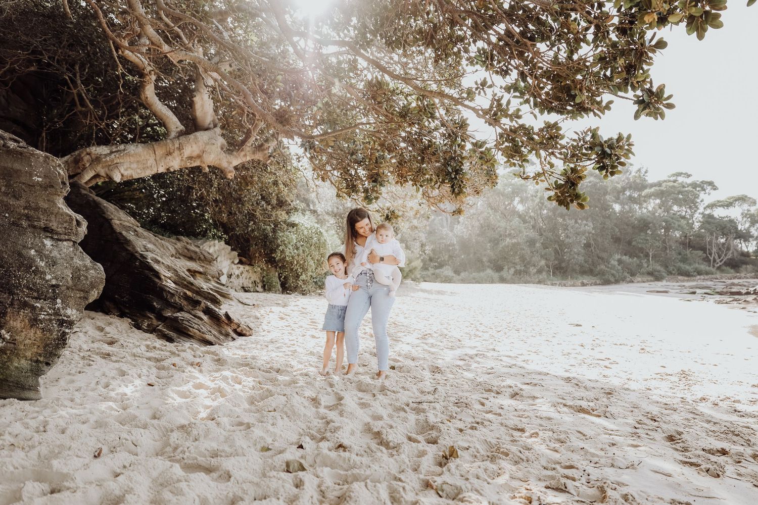 A mother holds her two children under a large tree on a bright sandy beach.