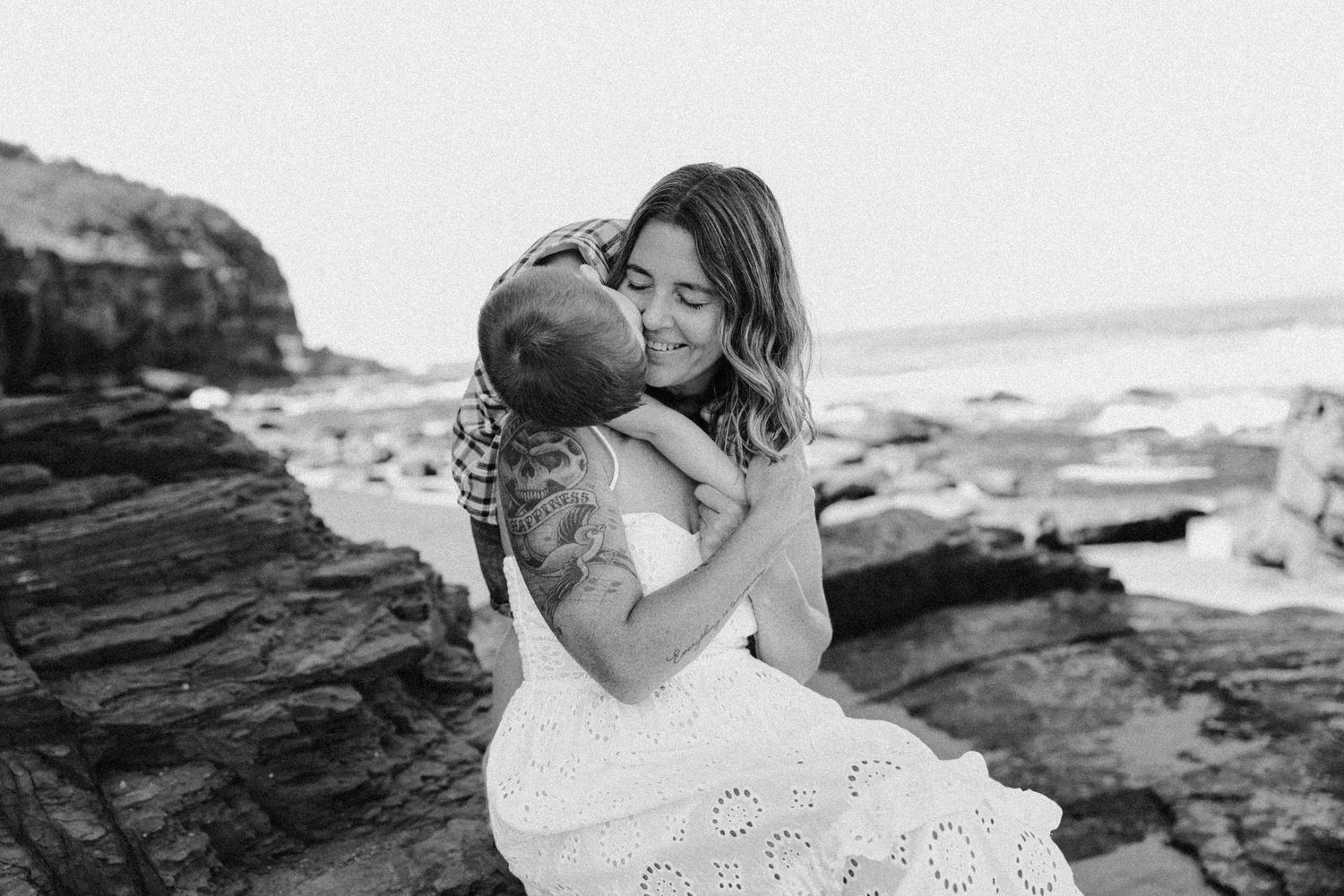 Black and white photo of a woman in lace dress embracing a child on rocky ocean shore.