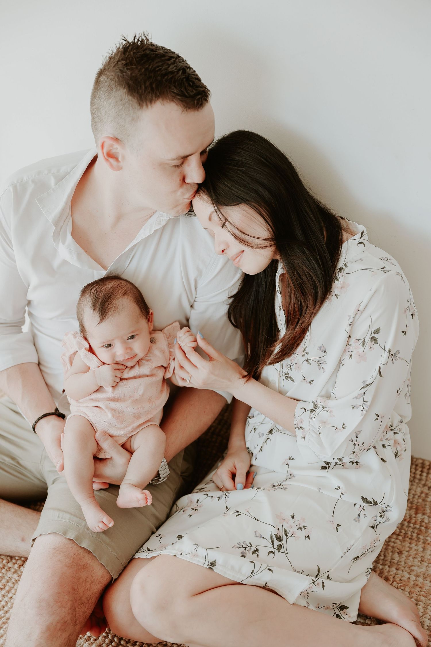 Young couple sitting on floor kissing over their newborn baby in a bright, airy room.