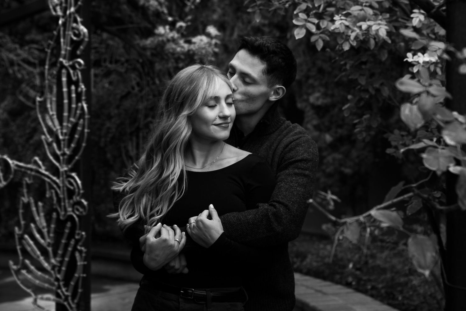 Black and white photo of a romantic couple embracing outdoors, man kissing woman's cheek among lush garden greenery.