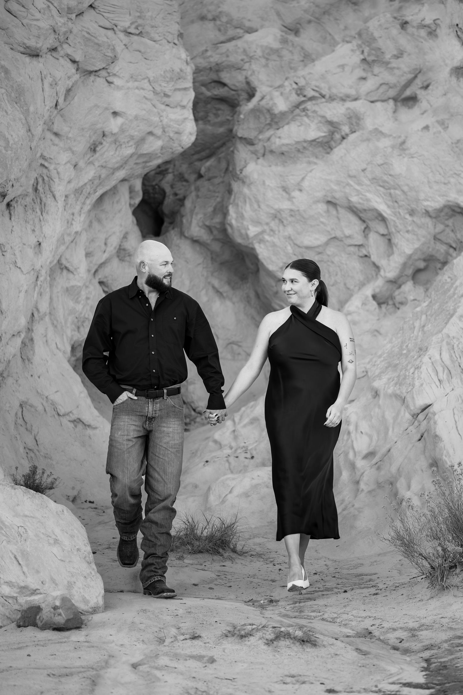 Black and white photo of a couple holding hands and walking through a rocky canyon, smiling at each other.