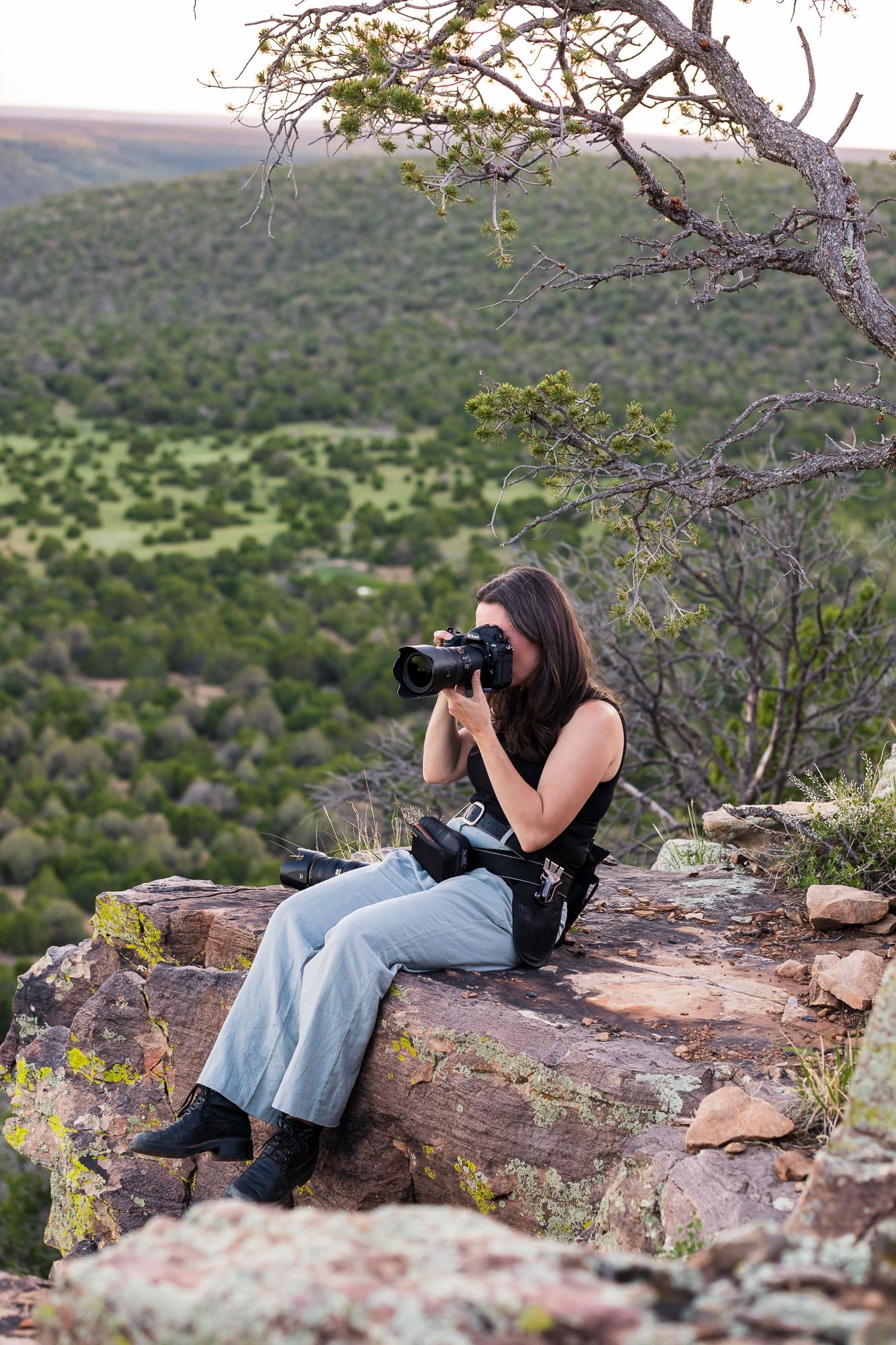 A photographer lies on a rocky cliff edge taking photos of the scenic mountain landscape below.