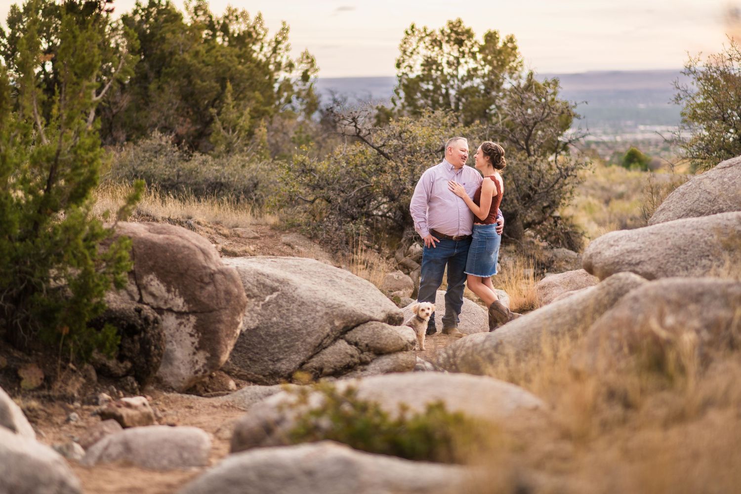 Couple embracing on rocky terrain surrounded by desert shrubs and scenic landscape at golden hour.