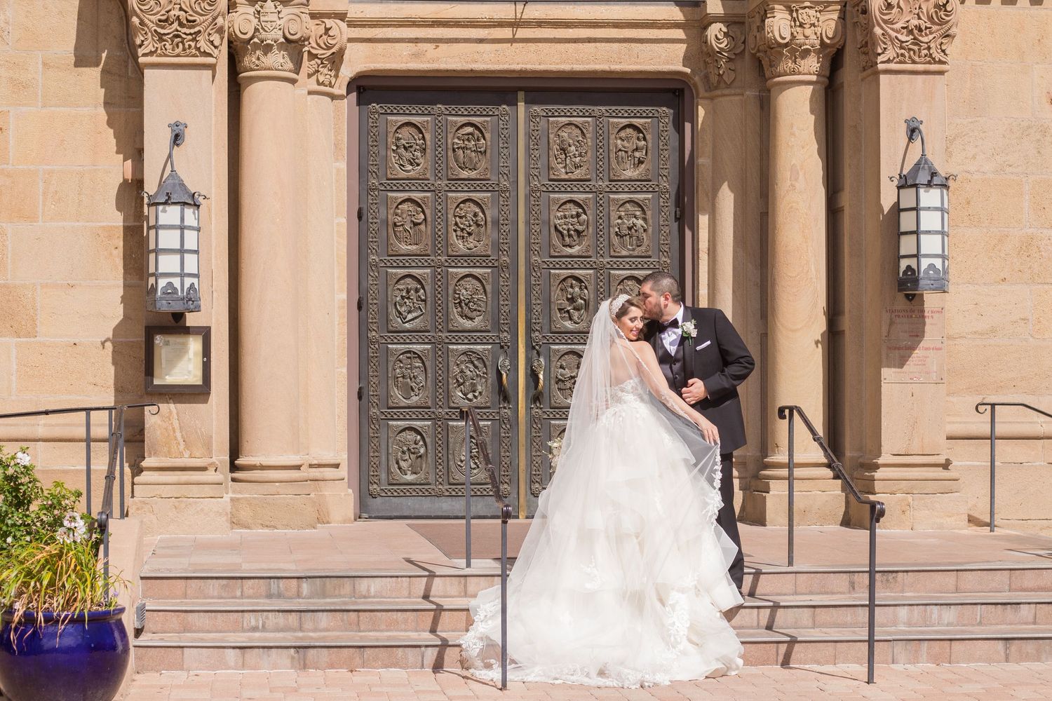 A couple shares a romantic moment on stone church steps in front of an ornate carved wooden door with lanterns.
