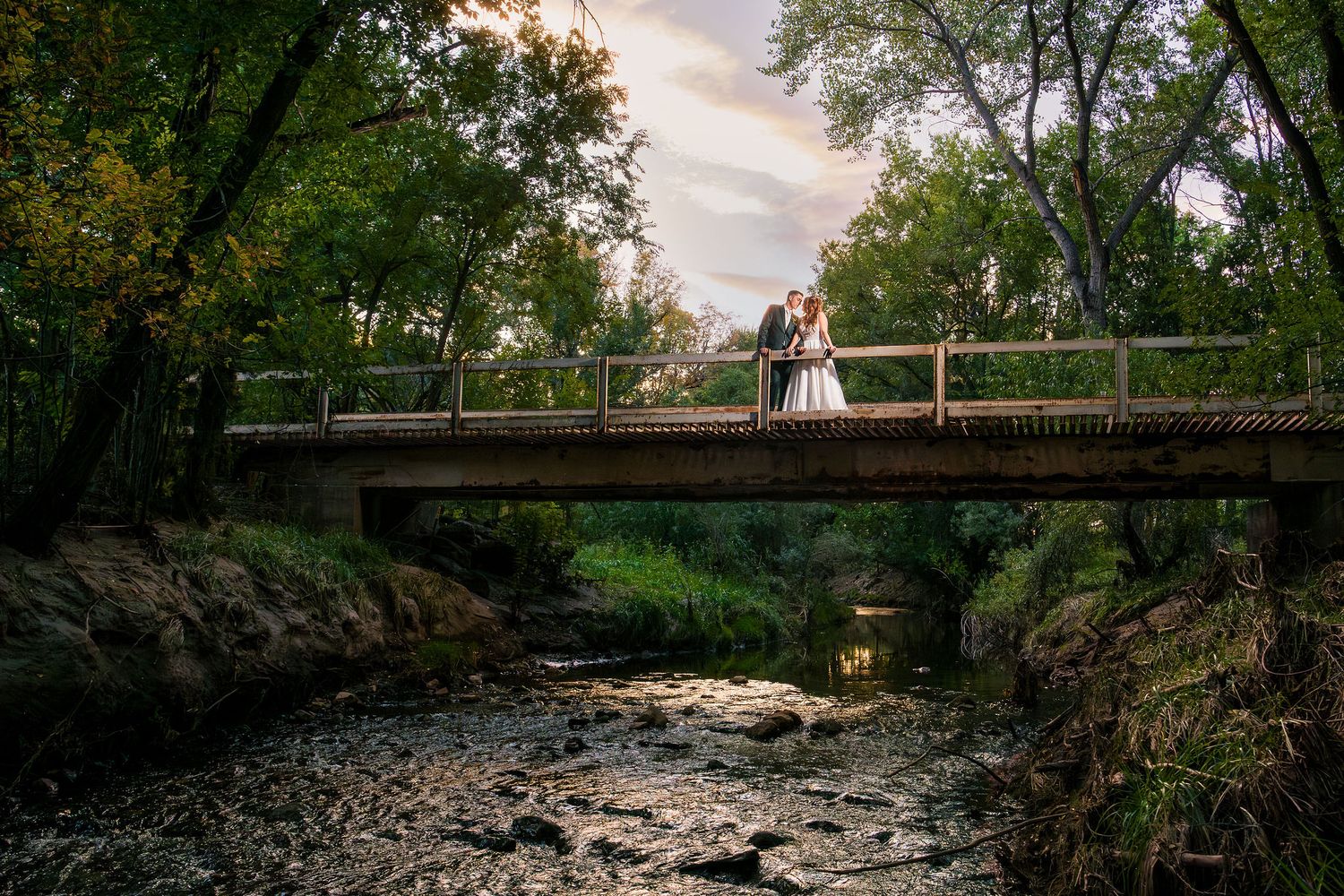 Bride and groom kissing on a bridge over a rocky stream surrounded by lush green trees at sunset at San Patricio Orchards in Ruidoso, NM..