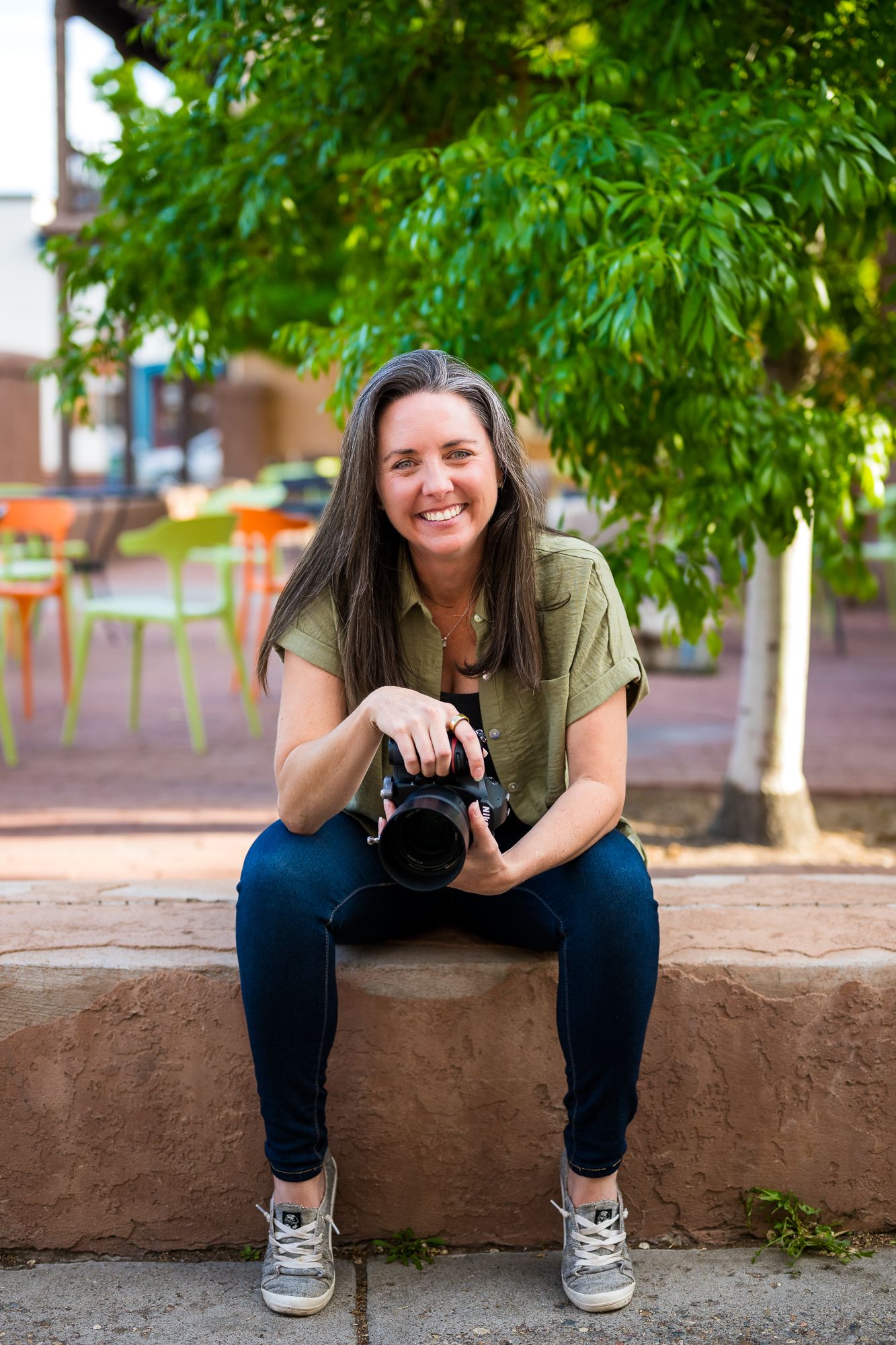 Smiling woman sitting outdoors holding a DSLR camera, wearing a green shirt and jeans, with trees in the background.
