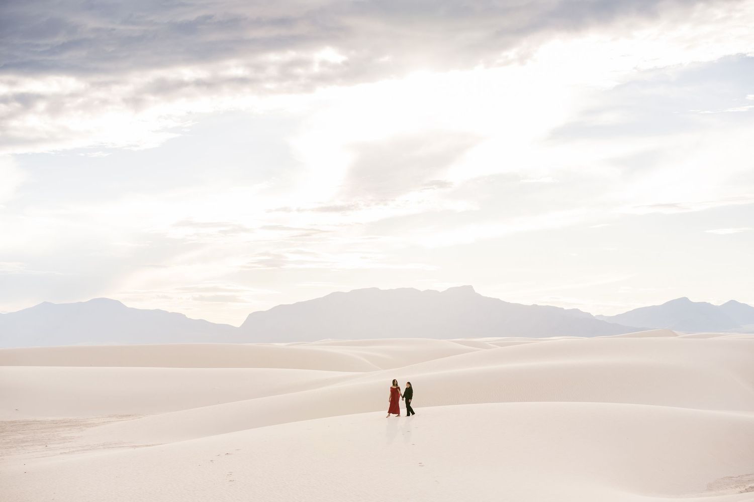 Two figures in colorful attire walk across vast white sand dunes with mountain backdrop at White Sands National Park.
