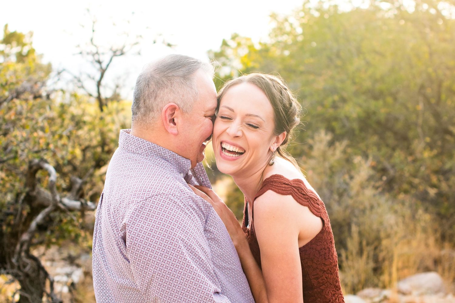 A laughing couple embraces outdoors in warm golden sunlight surrounded by natural desert vegetation.
