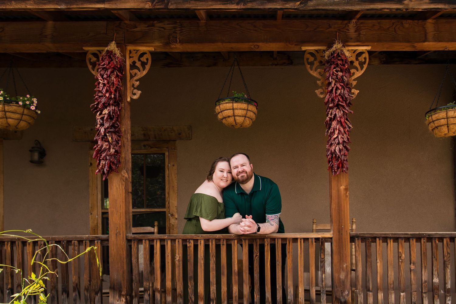 A couple stands together on a rustic porch decorated with hanging dried chili peppers and wooden beams.