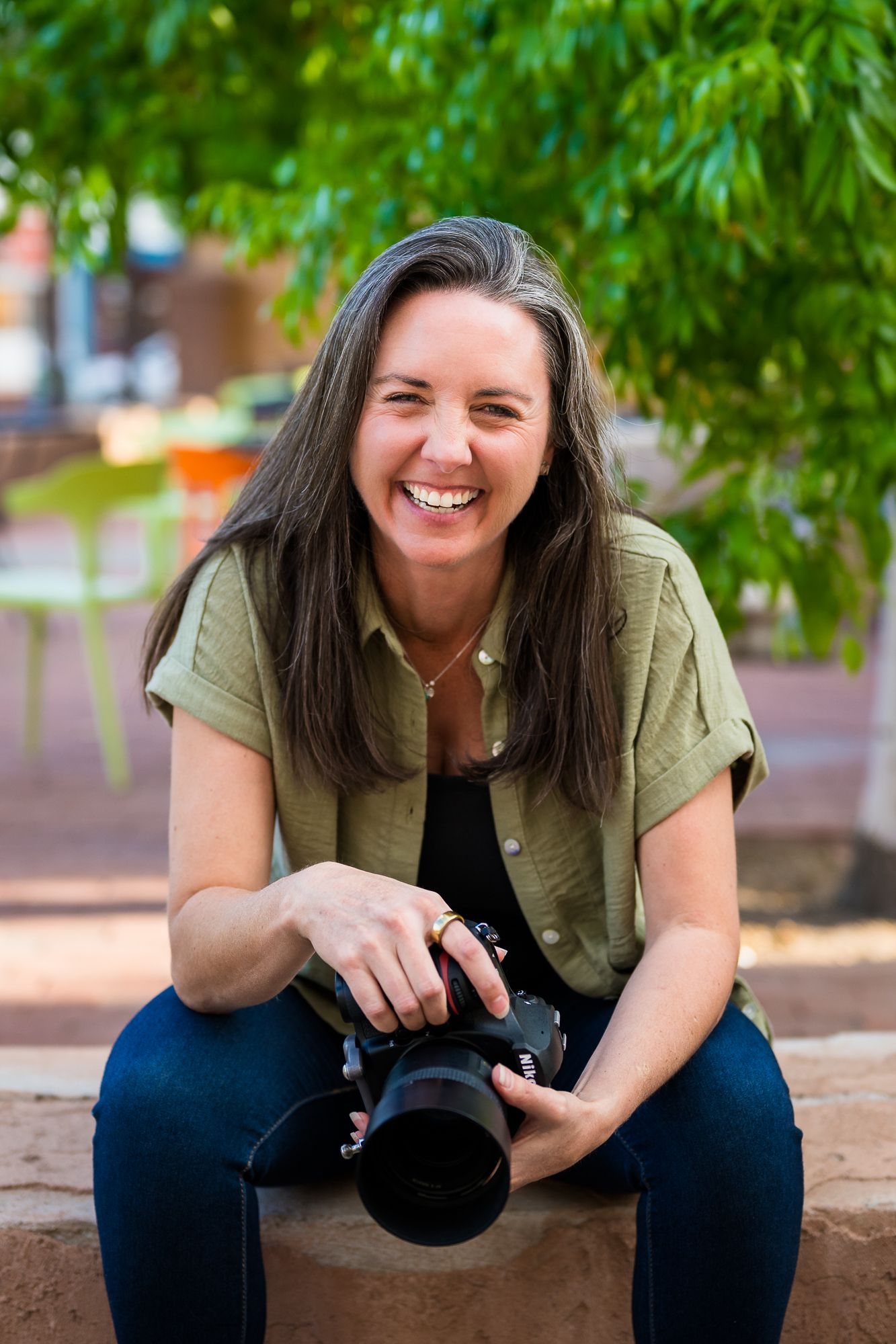 Smiling woman with long dark hair holding a DSLR camera, sitting outdoors with green trees in the background.