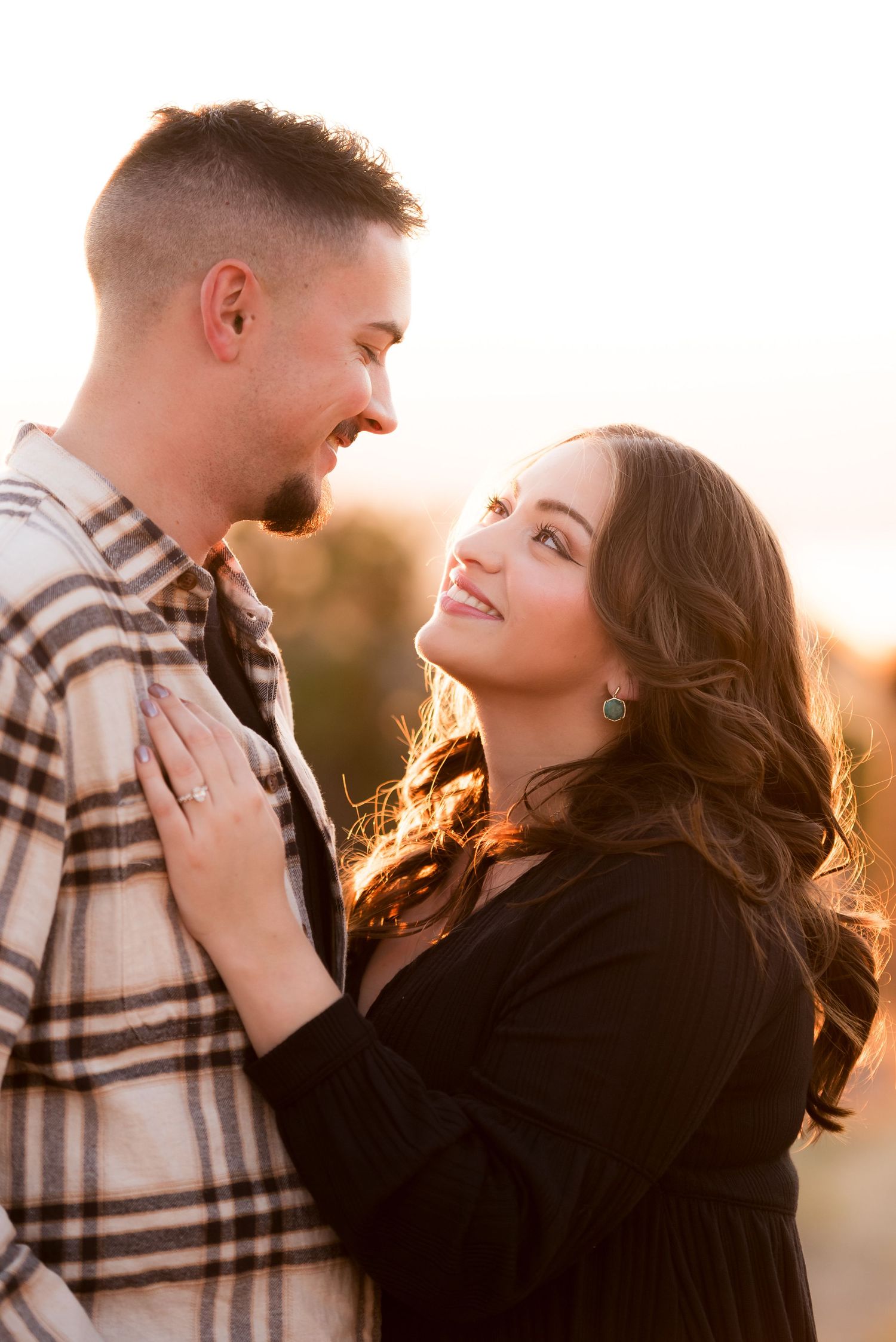 A couple shares a romantic golden hour moment outdoors, woman in black smiling up at man in plaid shirt.