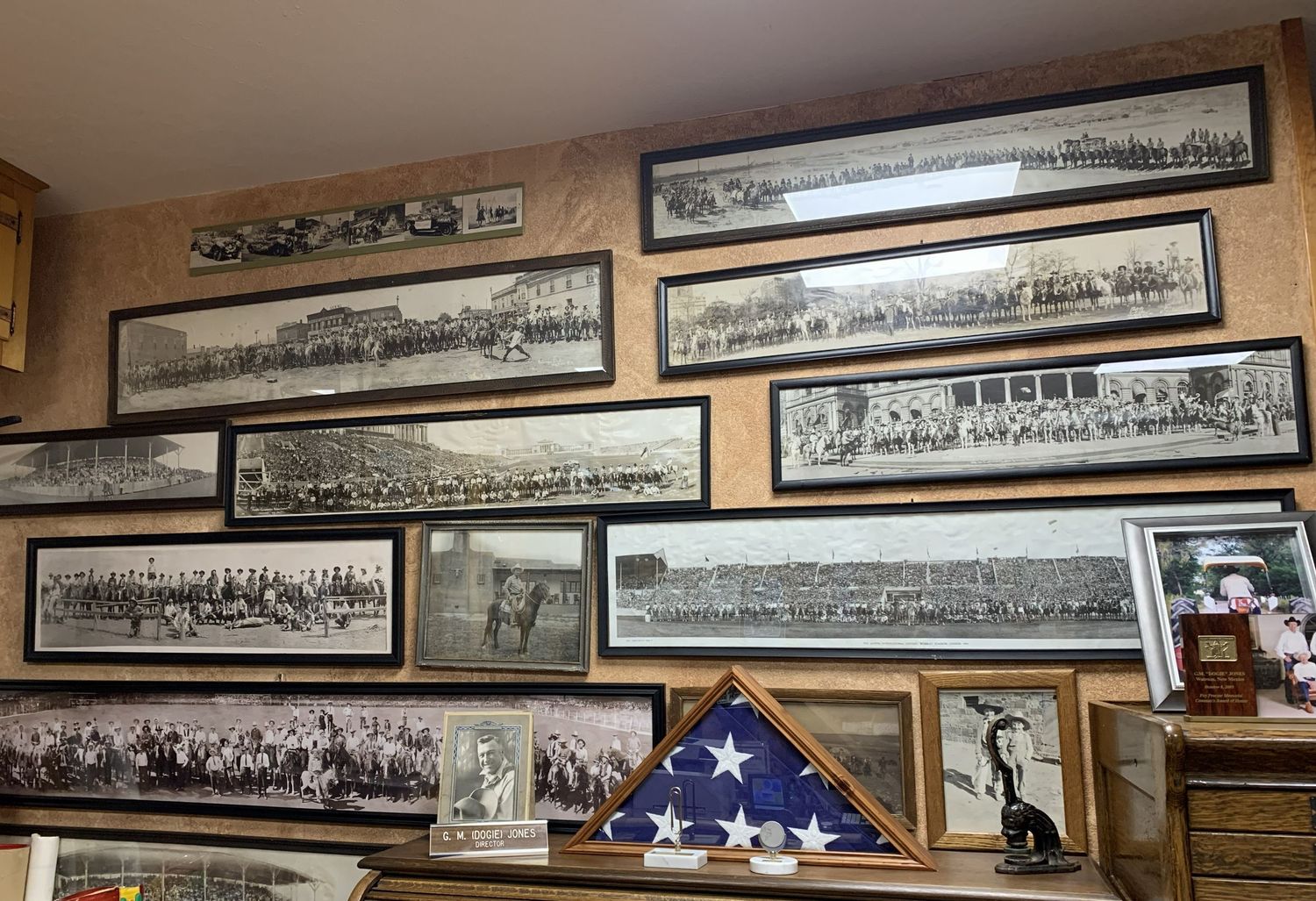 Vintage black and white panoramic photos displayed in frames on a wood-paneled wall above a folded flag in a case.
