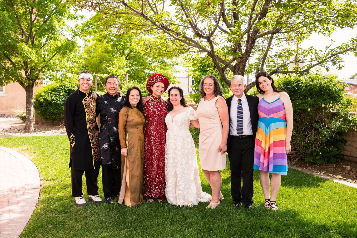 A group of formally dressed people stand together on a lawn with green trees in the background on a sunny day.