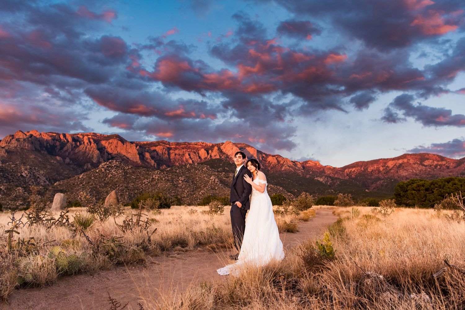 A couple stands in the desert landscape with dramatic red mountains and vibrant sunset clouds in the background.