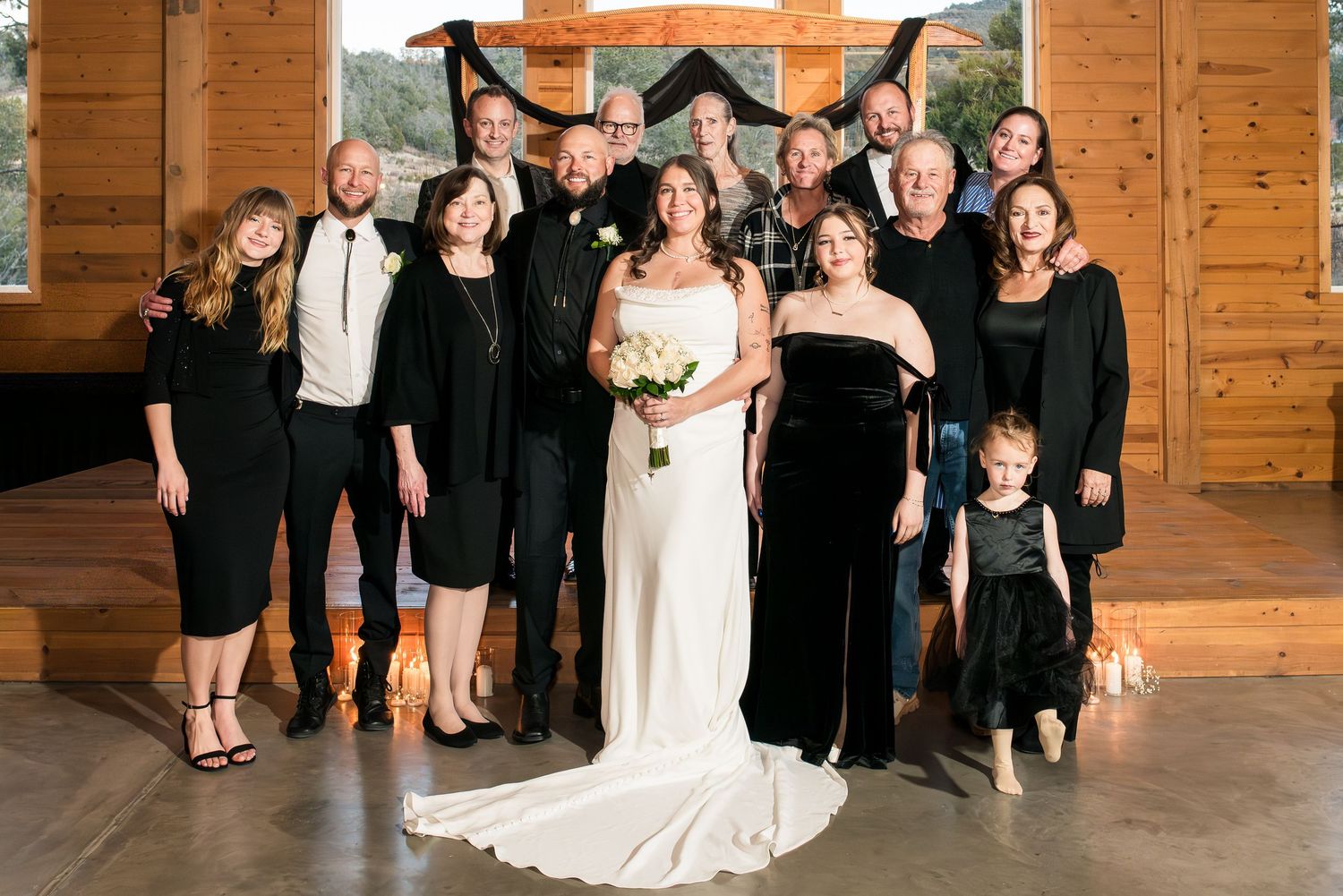 Wedding party members in formal black attire and white gowns pose together inside rustic wooden venue.