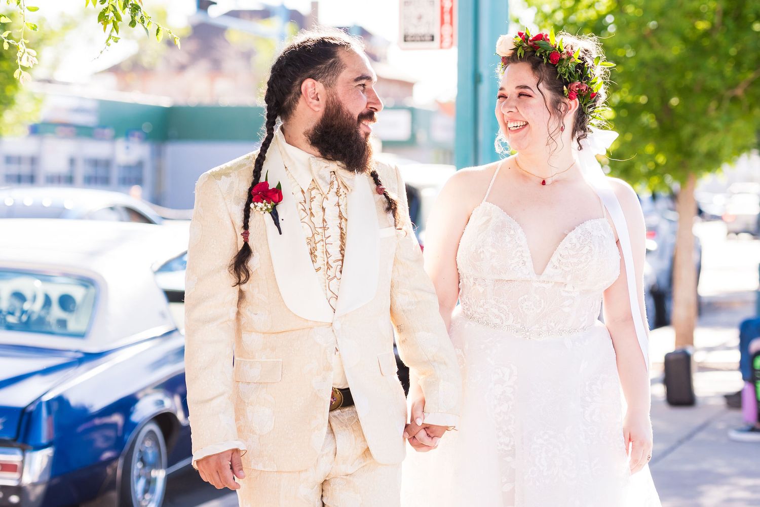 A smiling couple in wedding attire holds hands outdoors, groom in cream suit with braids, bride in lace dress with floral crown.