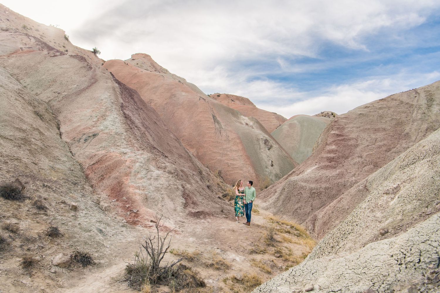 Hikers explore colorful striped badlands with pink and gray layered rock formations under cloudy skies.