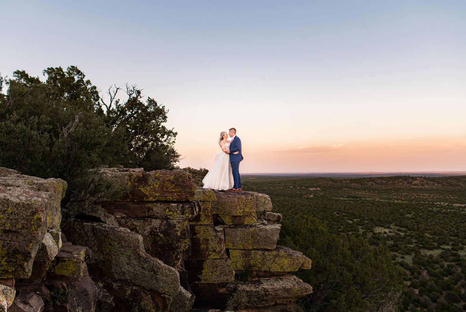 A couple stands silhouetted together on a rocky cliff overlooking a dramatic sunset landscape with atmospheric skies.