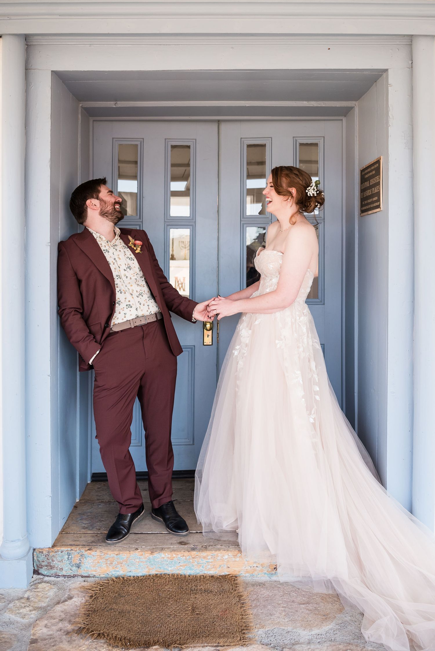 Bride in white gown and groom in burgundy suit hold hands smiling at each other before a blue double doors at Bishops Lodge in Santa Fe.