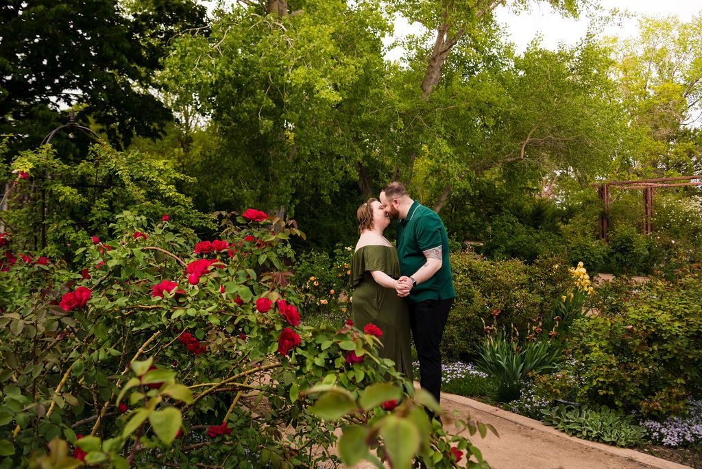 Couple sharing a romantic kiss surrounded by lush greenery and red roses in a beautiful garden setting.