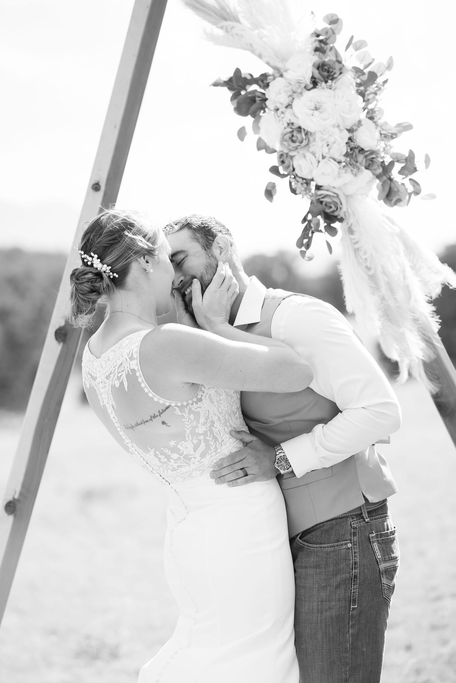 Black and white photo of a bride and groom sharing their first kiss under a floral arch at an outdoor wedding.