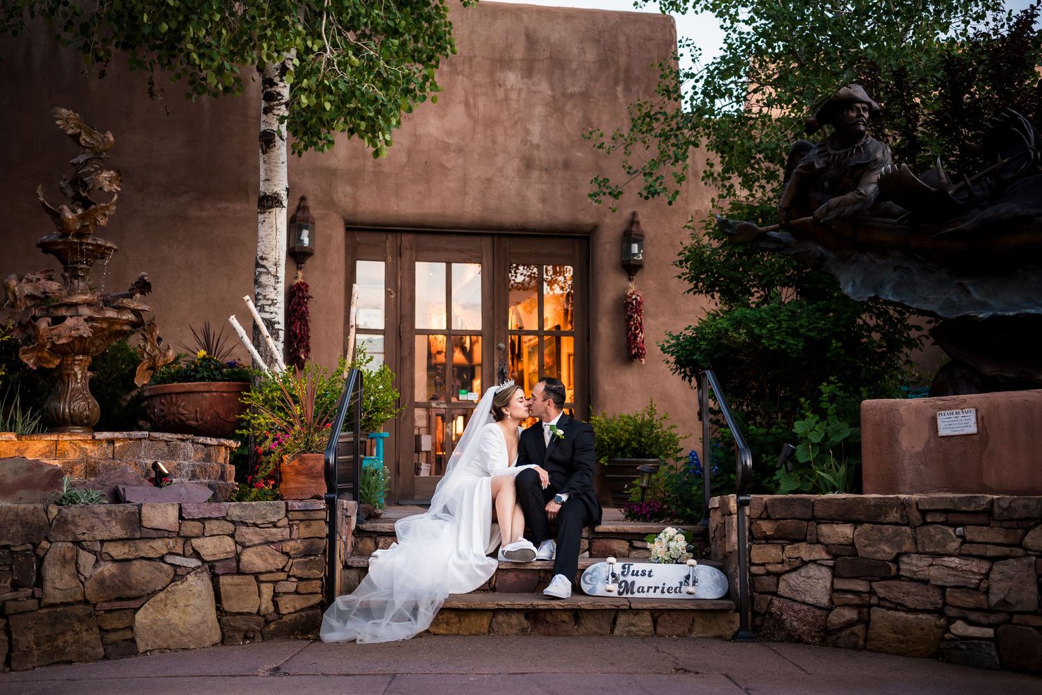 A romantic wedding couple poses in front of an adobe-style building at sunset with string lights overhead.