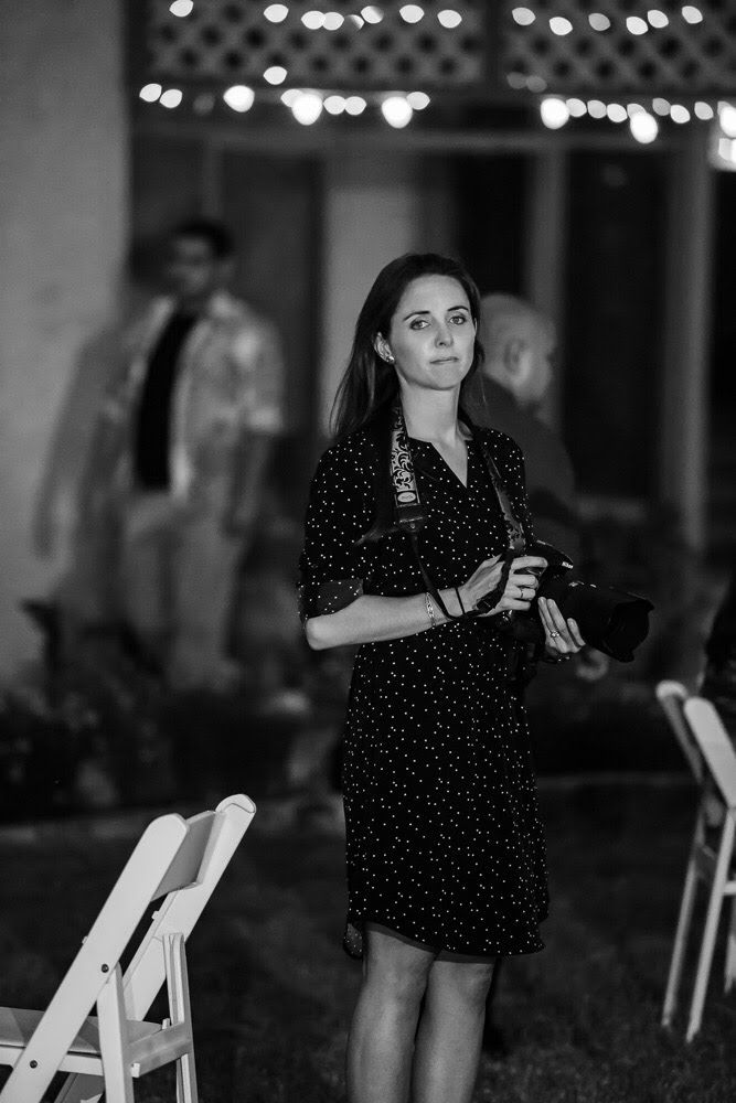 Black and white photo of a woman in a polka dot dress holding a camera at an outdoor evening event.