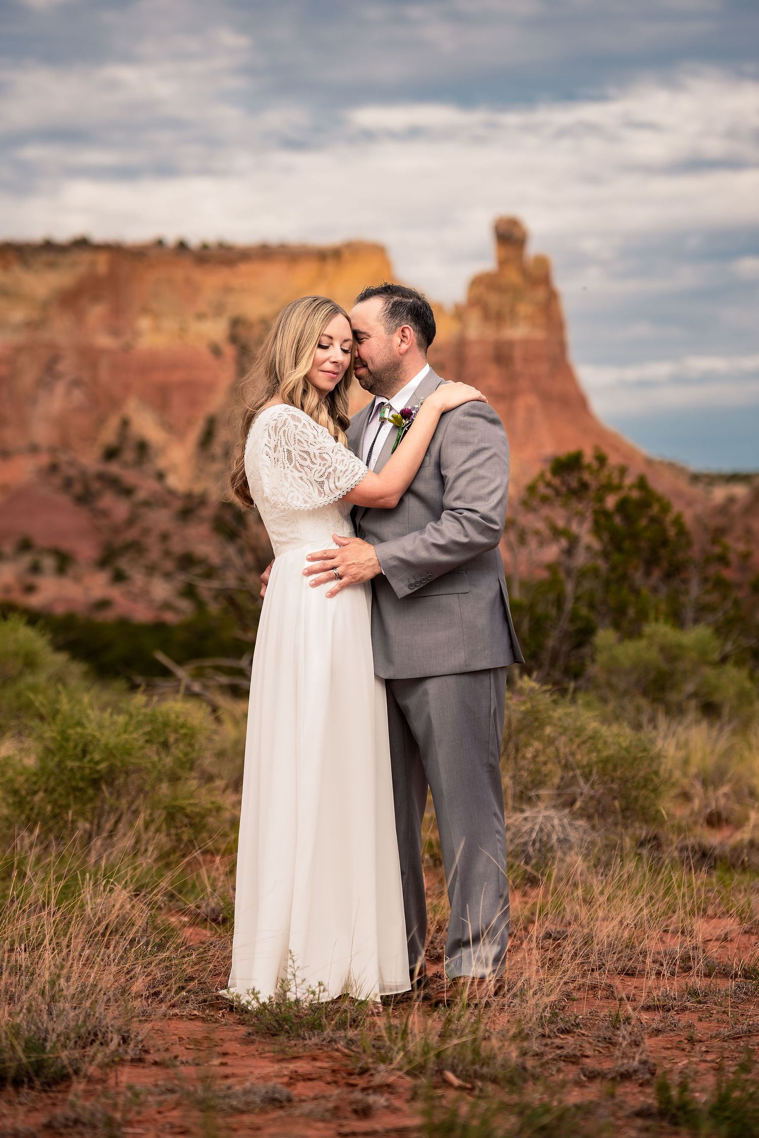 Couple embracing in wedding attire with red rock formations and desert landscape in the background.