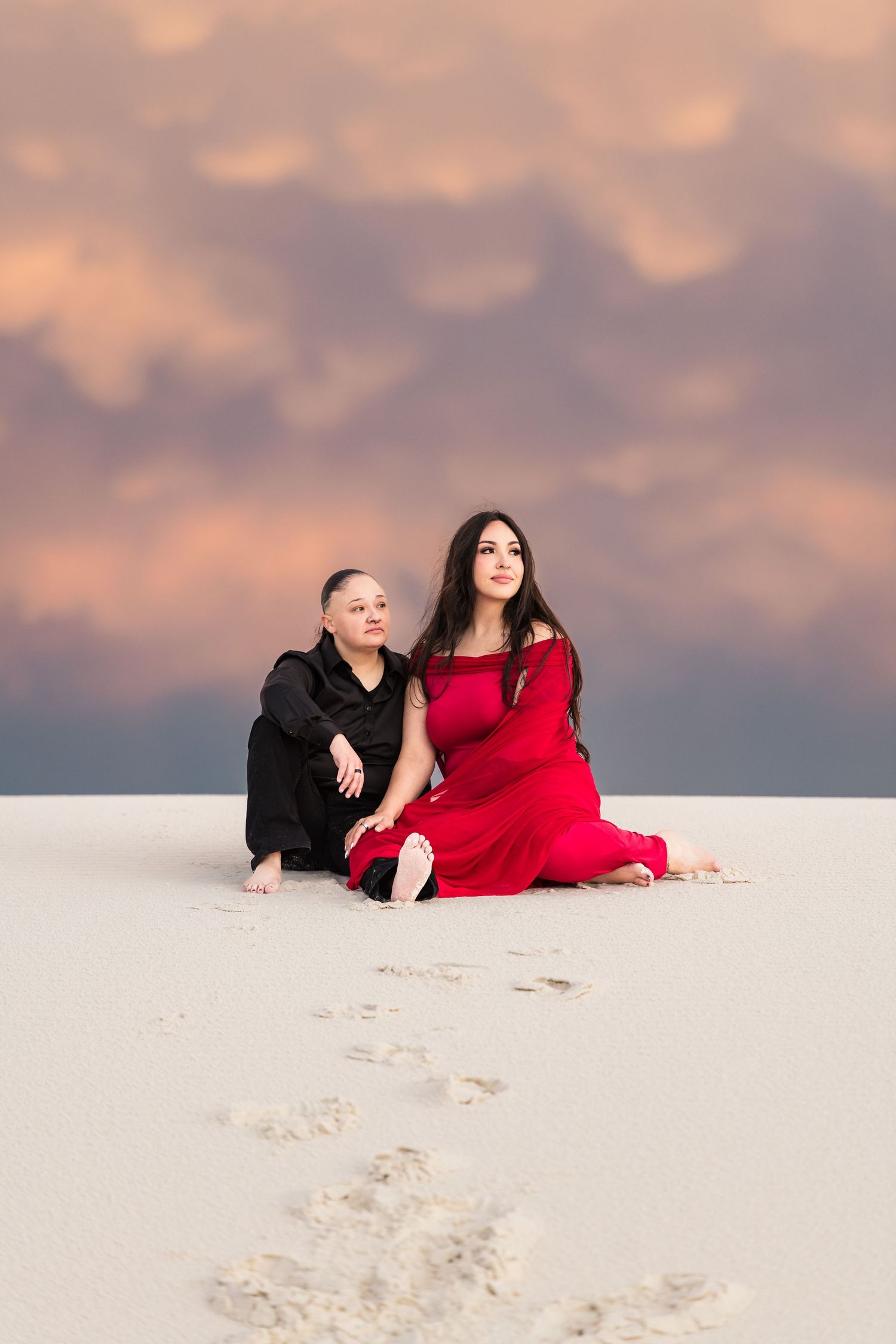 Two women in red and black outfits sit barefoot on white sand dunes under a dramatic cloudy sunset sky.