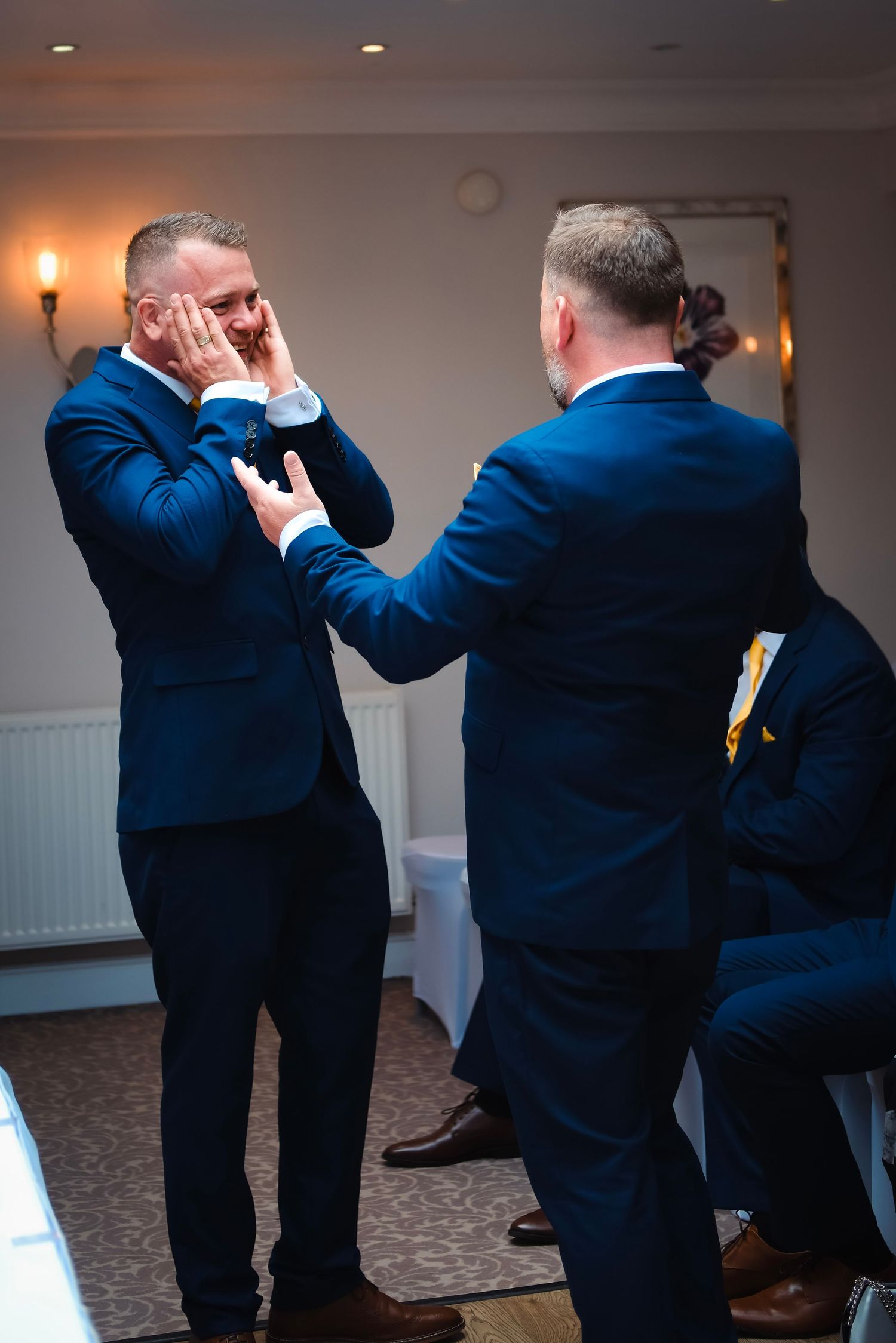 Two men in navy suits sharing an emotional moment during wedding preparations.