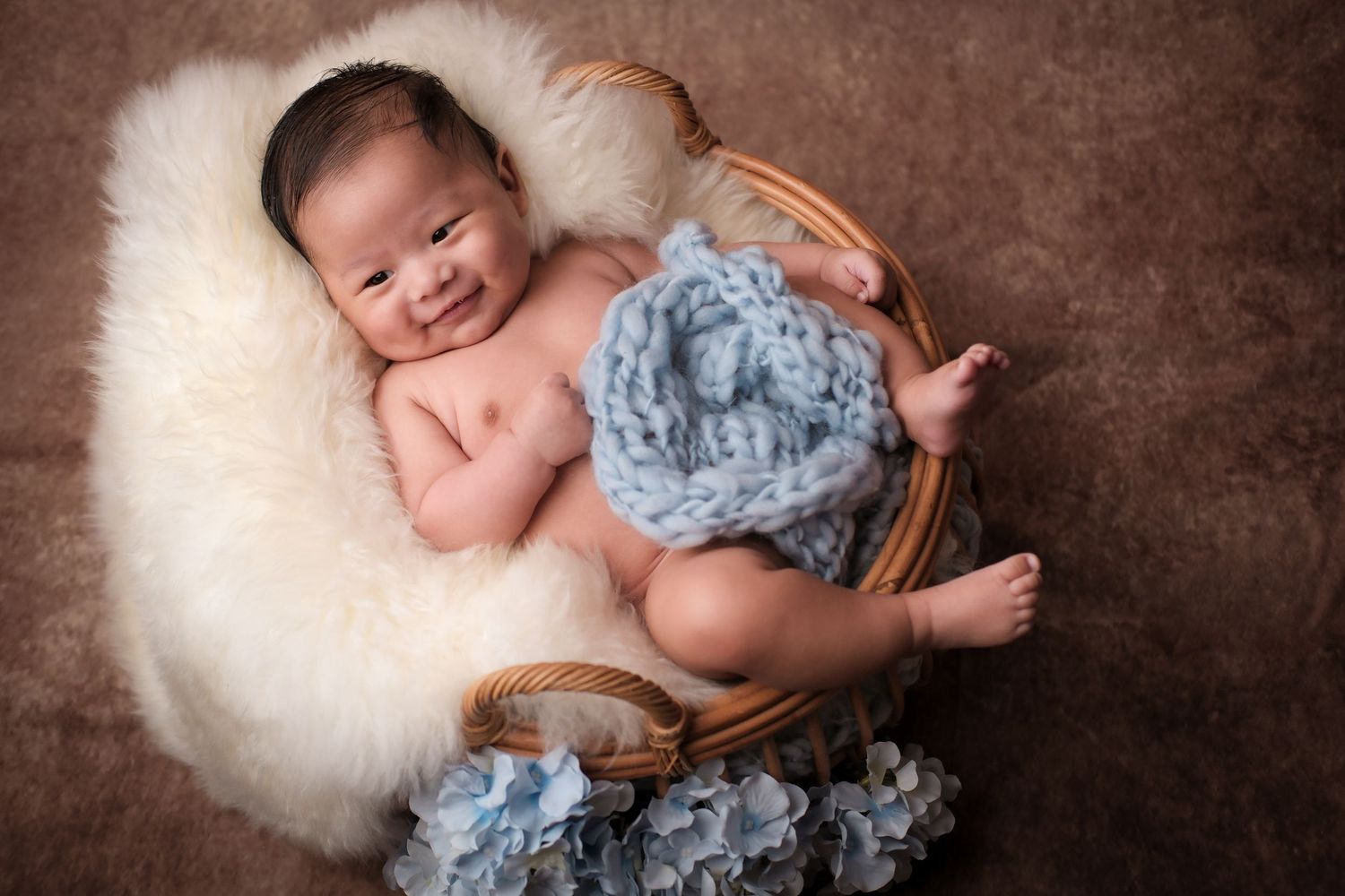 Sleeping newborn nestled in basket with blue floral decorations on white fur backdrop.