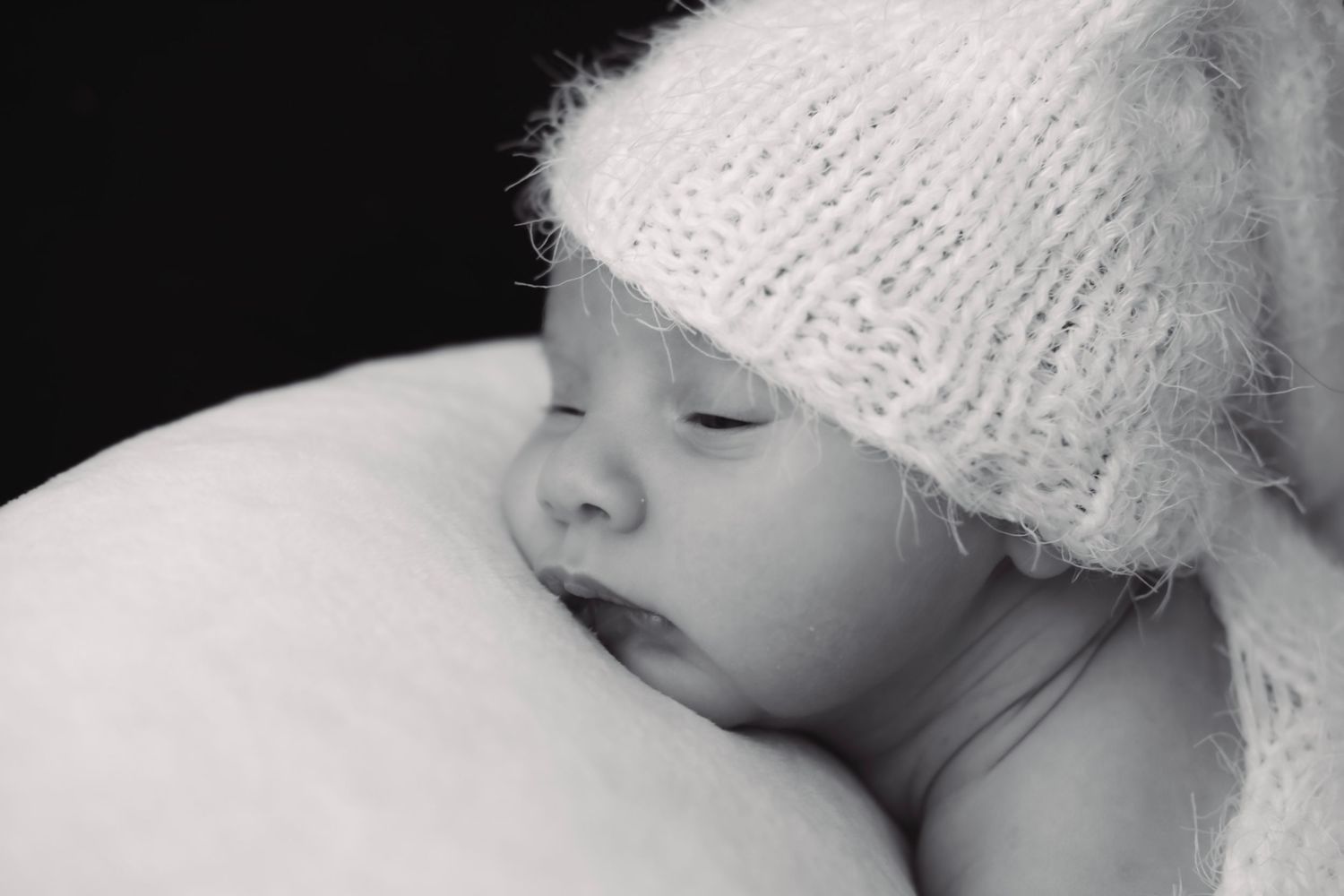 Sleeping newborn wearing white knit hat rests on soft pillow in black and white portrait.