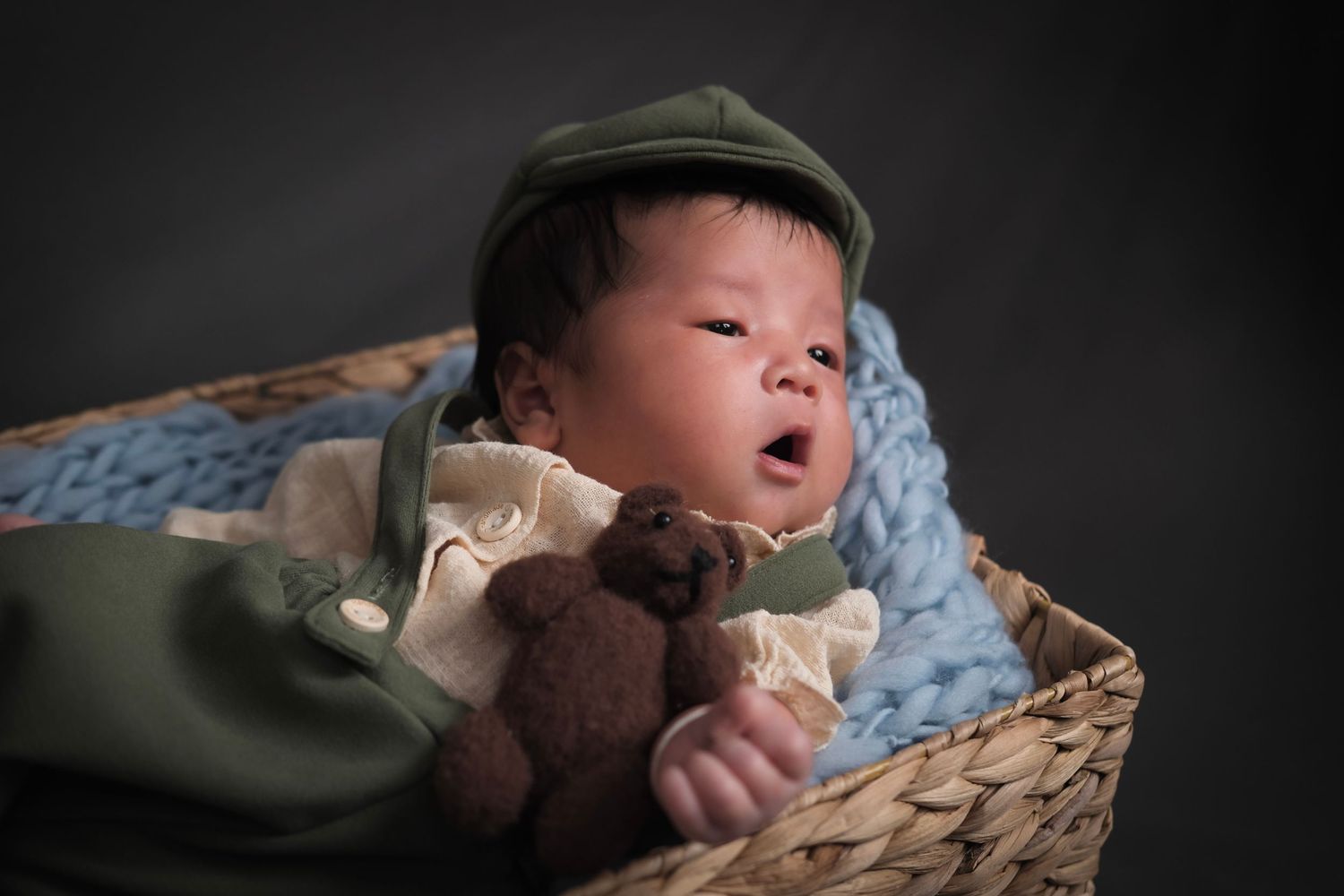 Newborn baby sleeps in woven basket with blue blanket against dark backdrop.
