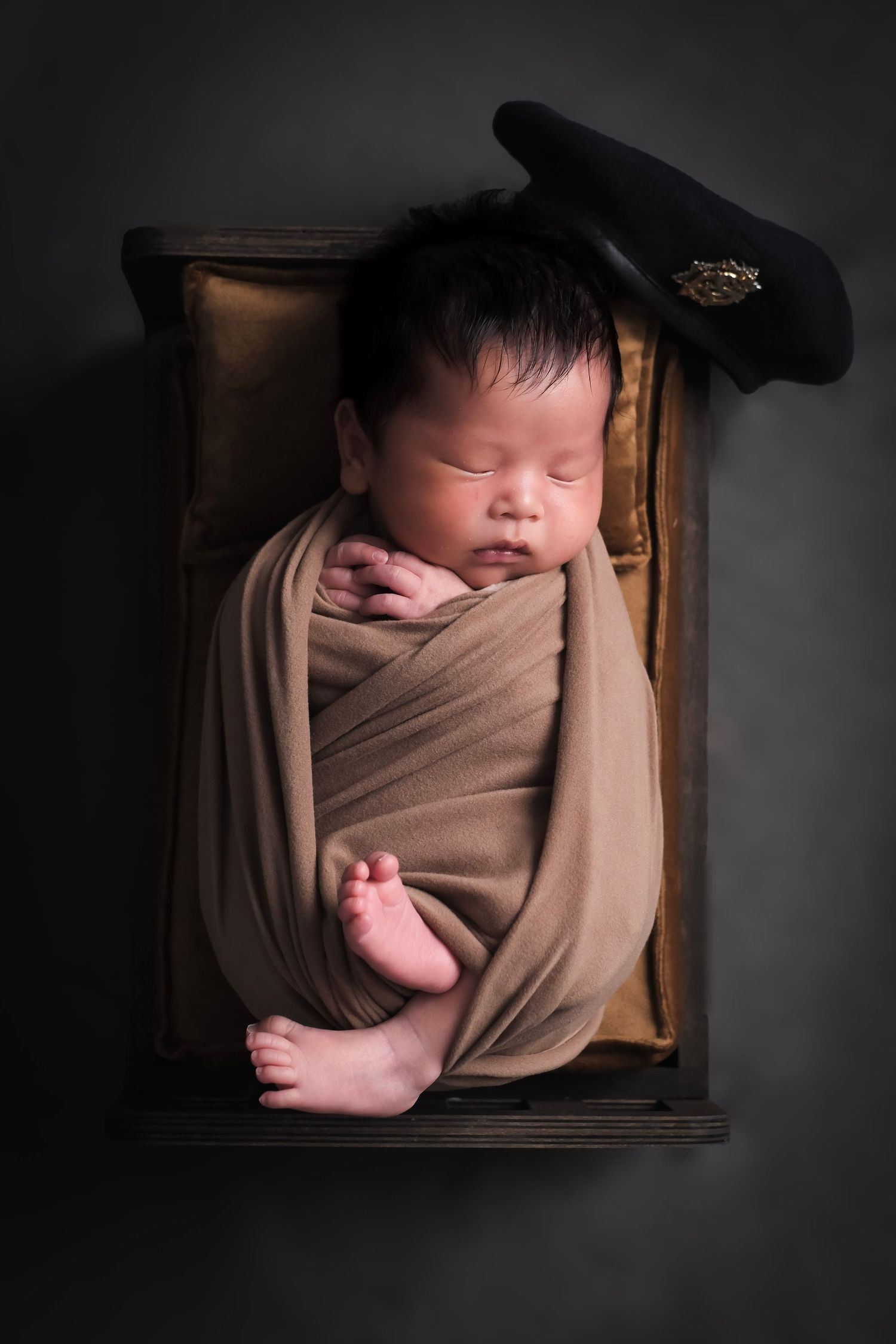 Sleeping newborn wrapped in tan fabric with police cap in dark wooden box.