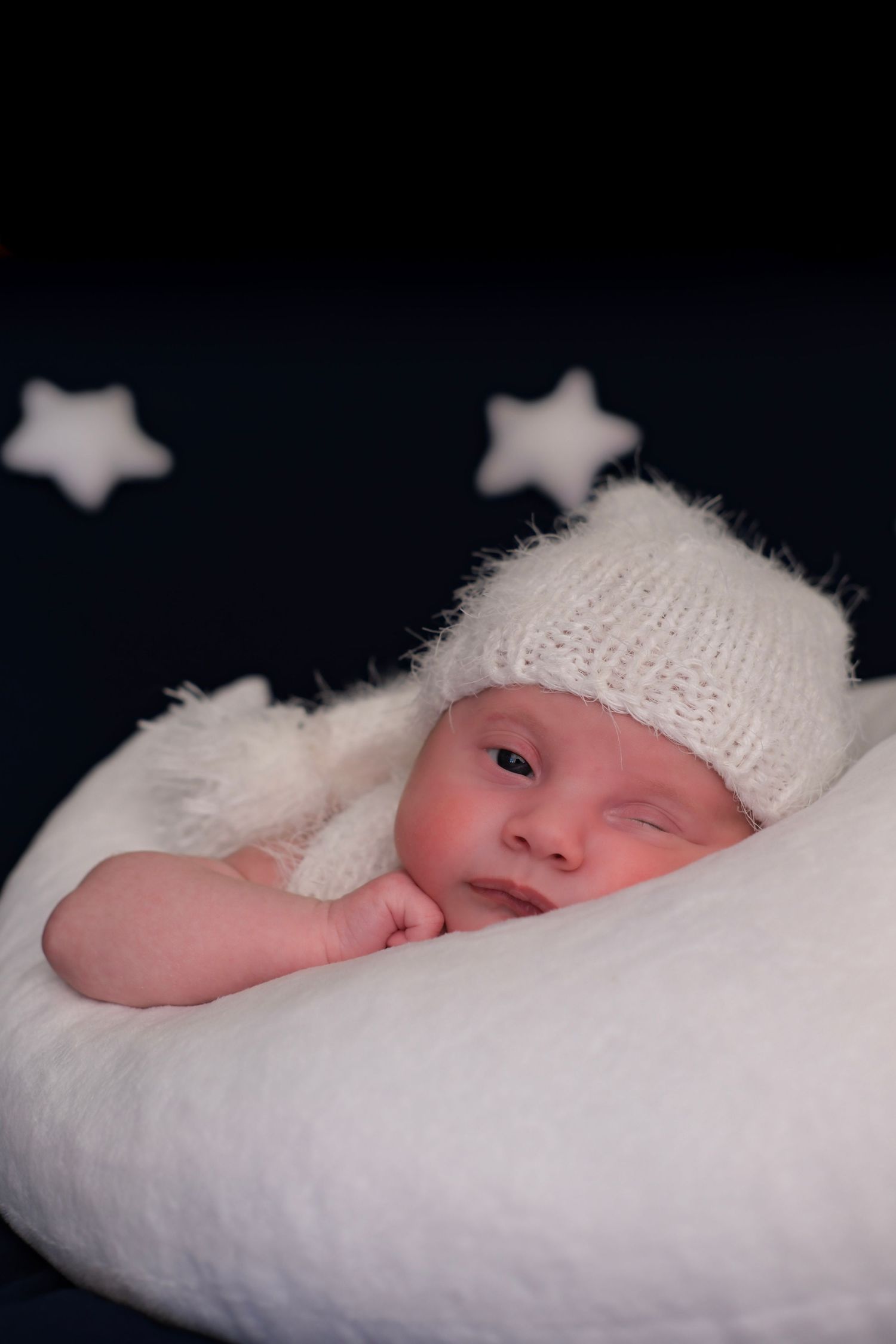 Newborn in white knit hat rests on pillow with star decorations in background.