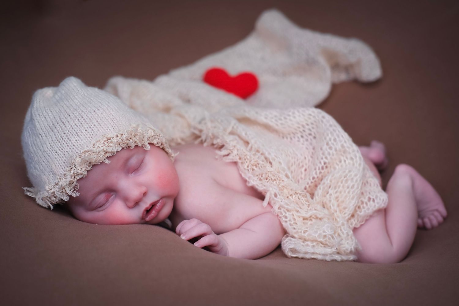Sleeping newborn covered in delicate lace fabric with small red heart accent.