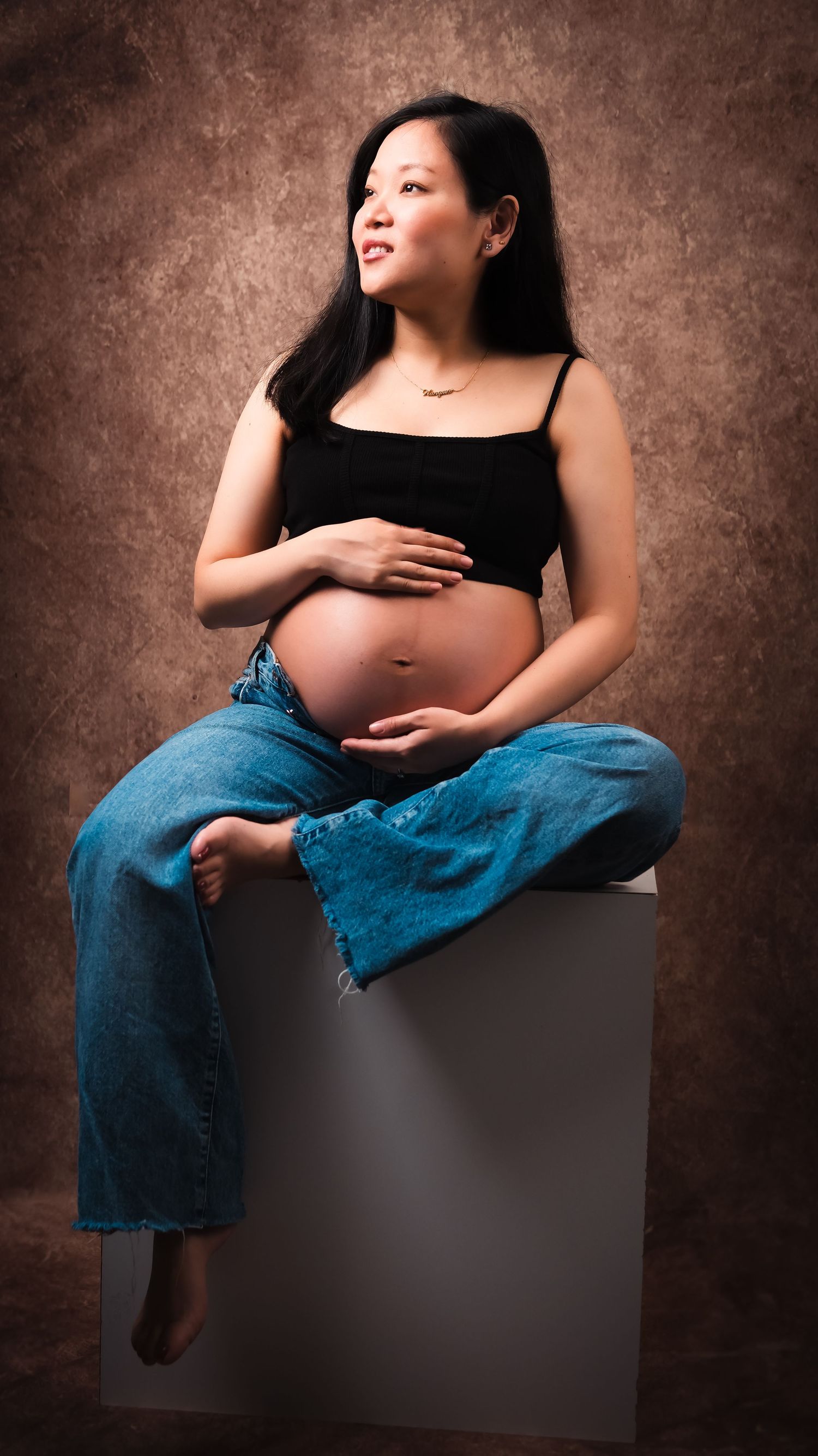 Expectant mother in casual denim and black top poses during studio maternity session.