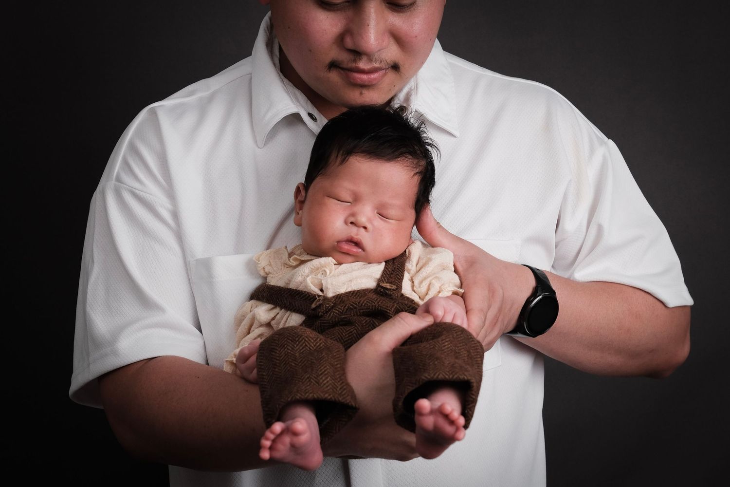 Newborn baby wearing a brown outfit being cradled in studio lighting.