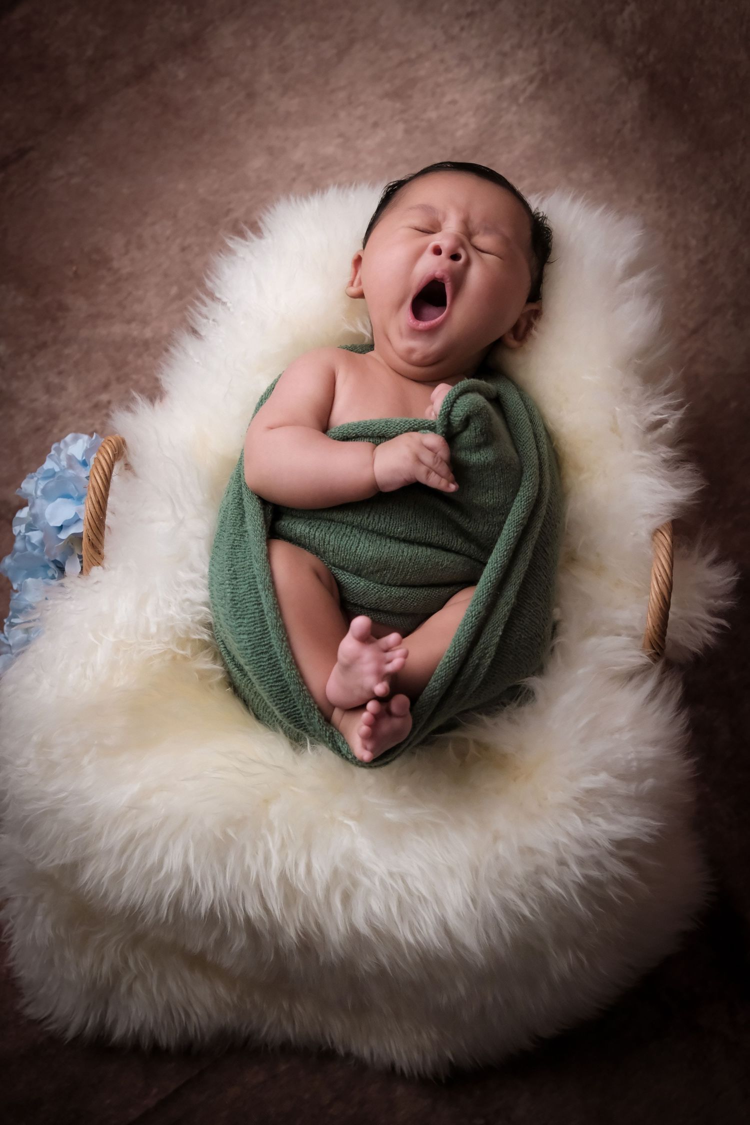 Newborn baby wrapped in green fabric yawns while lying on soft white fur.
